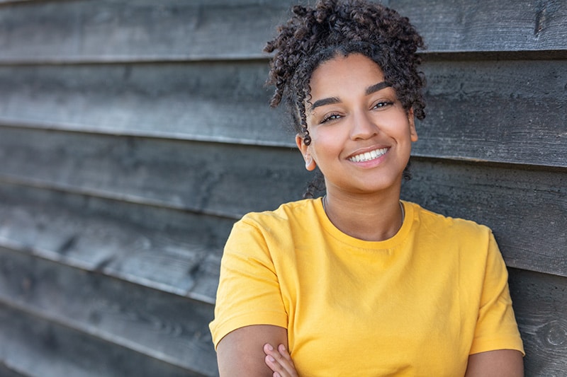 Smiling young woman with curly hair wearing a yellow shirt against a wood plank backdrop in the homepage header at Advanced Orthodontics in Everett, WA